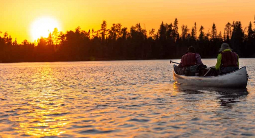 two people sit in a canoe on calm water while the sun sets behind trees on the shore